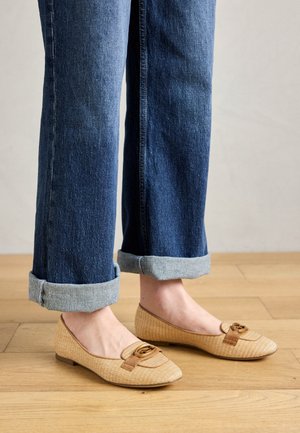 Ballerines beiges tissées avec un bout carré, des accents bruns et un détail de boucle en or. Associées à un jean en denim noir avec les ourlets retroussés sur un sol en bois.