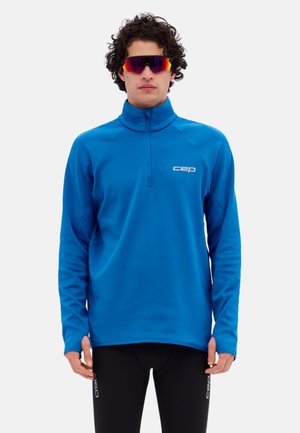 Mann trägt ein blaues Sportoberteil mit Reißverschluss und langen Ärmeln sowie schwarze sportliche Leggings, mit reflektierenden Sportsonnenbrillen.