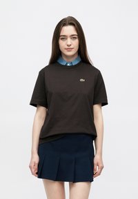 Pruun puuvillane T-särk ümara kaelusega ja lühikeste varrukatega, millel on väike roheline Lacoste'i logo. Sobitatud tumesinise volditud seelikuga.