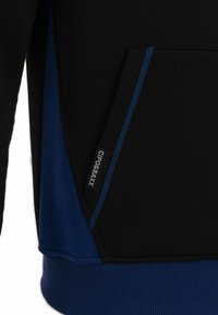 Sweatshirt noir et bleu avec un tissu texturé, une poche latérale avec des coutures visibles, et une étiquette logo sur la couture inférieure.