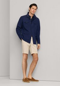 Man draagt een marineblauw jack, donker shirt, beige korte broek en bruine loafers, staand tegen een effen lichtgrijze muur.