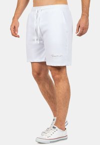 Witte zwembroek met een elastische tailleband, trekkoord, zijzakken en een geborduurd logo op het linkerpunt. Glad stoffen textuur.
