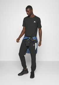 Svart atletisk t-shirt med vitt logotyp, matchande joggingsbyxor, lager med en blå jacka knuten runt midjan, och svarta sneakers.