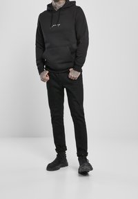 Schwarzer Hoodie mit einer Fronttasche und weißem Textlogo, kombiniert mit schwarzen Slim-Fit-Jeans und schwarzen Sneakers. Glatte, matte Textur.