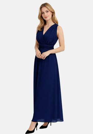 Kobieta o blond włosach, ubrana w granatową, bezrękawnikową suknię do ziemi oraz czarne wysokie obcasy, stojąca na tle jednolitego, jasnego tła.