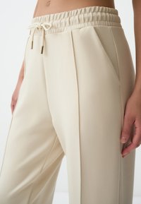 Beige joggerbroek met een elastische tailleband, aantrekkoord met gouden accenten en zijzakken; gemaakt van zachte, gladde stof met plooien.