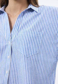 Camicia a righe blu e bianche con colletto e taschino sul petto, realizzata in tessuto leggero; presenta righe verticali di larghezze variabili.