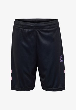 Kind draagt zwarte sportshorts met paarse logo en pijlen, zwarte sokken en witte sneakers met blauwe chevrons, staand op een effen achtergrond.