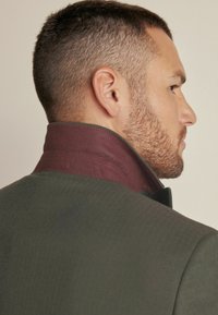 Olive green jacket featuring a high collar with a burgundy accent. The texture appears smooth with subtle pattern details.