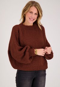 Bruine gebreide trui met een geribbelde textuur, oversized mouwen en een ronde halslijn, gecombineerd met donkere spijkerbroek en gouden armbandaccenten.