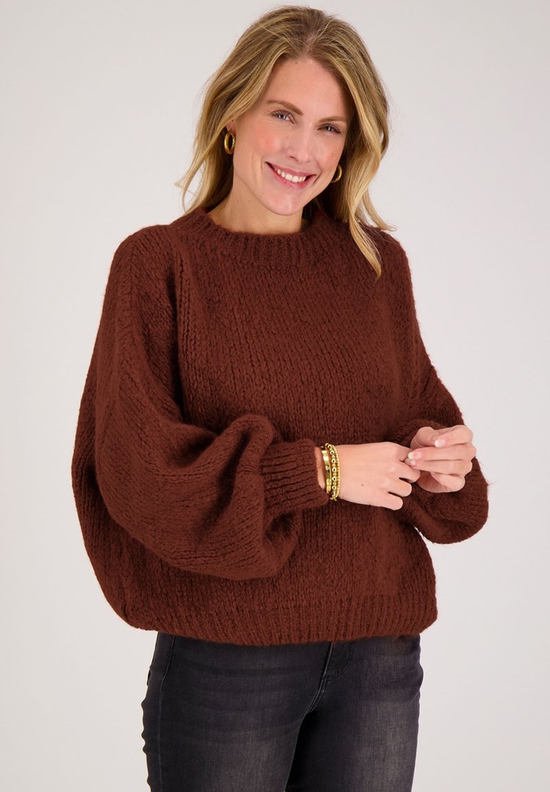 Bruine gebreide trui met een geribbelde textuur, oversized mouwen en een ronde halslijn, gecombineerd met donkere spijkerbroek en gouden armbandaccenten.