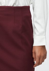 Desna ruka odmara se uz visoko strukiranu burgundsku suknju, koja se nosi s bijelom majicom dugih rukava.