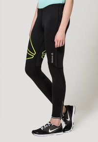 Schwarze Sportleggings mit gelben Akzenten, aus dehnbarem Material, mit einer Reißverschlusstasche und Reebok-Logo an der Seite.