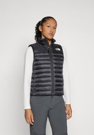 Black insulated vest with a high collar, quilted design, and front zipper. Features a logo on the left chest area. Worn over a long-sleeve shirt.