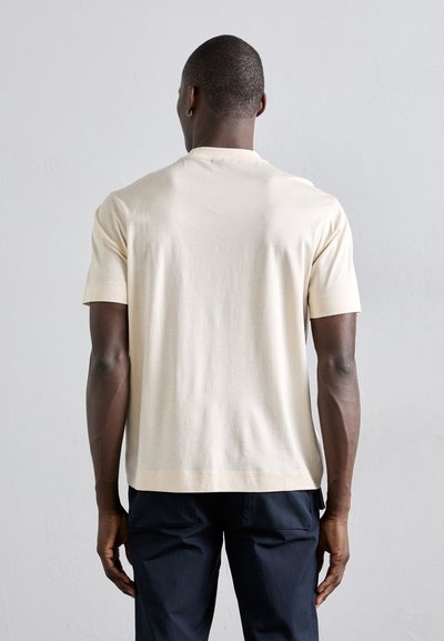 Helles beiges T-Shirt mit kurzen Ärmeln und rundem Ausschnitt, mit glatter Textur, kombiniert mit dunklen Hosen, von hinten betrachtet.