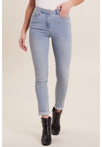 Jeans skinny bleu clair avec taille haute, design cinq poches et ourlets roulés, associés à des bottines noires. La texture en denim est visible.