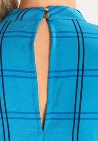 Top turchese con apertura a chiave sul retro, chiusura con bottone e motivo a quadri blu scuro su tessuto liscio.