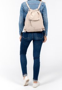 Mochila de cordón beige con un acabado texturizado, correas ajustables y un bolsillo frontal con cremallera, llevada con una chaqueta de mezclilla y jeans.