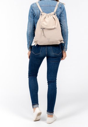Zaino a drawstring beige con finitura testurizzata, spallacci regolabili e tasca frontale con zip, indossato con una giacca di jeans e jeans.