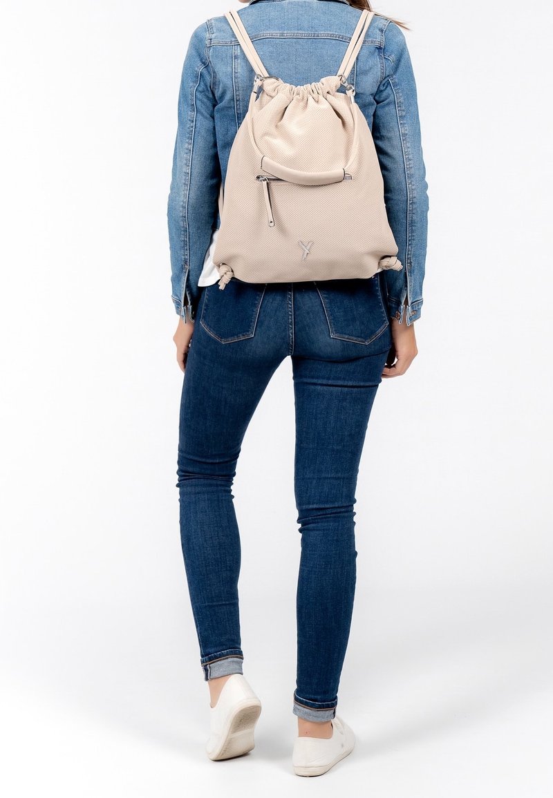 Mochila de cordón beige con un acabado texturizado, correas ajustables y un bolsillo frontal con cremallera, llevada con una chaqueta de mezclilla y jeans.