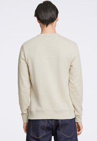 Felpa a girocollo beige chiaro con maniche lunghe, dotata di polsini e orlo a coste, e una texture liscia. Indossata sopra dei jeans scuri.