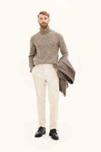 Grå stickad polotröja, beige byxor, svarta läderskor, håller en brun rutig jacka. Enkel, klassisk design med neutrala toner.