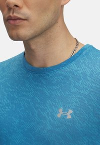 Camiseta deportiva azul con un patrón de punto texturizado, escote redondo y un pequeño logo plateado en el pecho. Collares de cadena visibles.