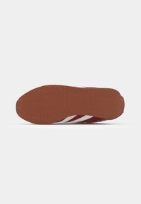 Red sports shoe with a textured brown rubber sole, featuring white accents and a herringbone tread pattern for grip.