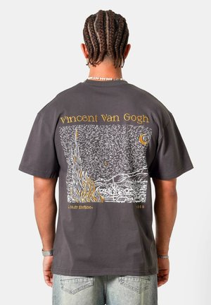 Person med flettet hår iført en mørkegrå T-shirt med stregtegning af Van Goghs maleri Stjernenat og teksten "Vincent Van Gogh".