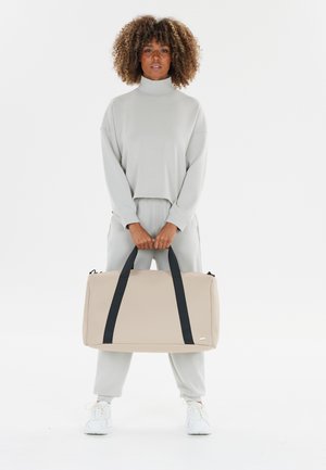 Woman with curly hair in light gray tracksuit holding beige duffel bag with black straps, standing against a plain white background.
