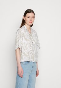 Camisa de manga curta, com botões, em bege com padrão botânico branco. O tecido tem uma textura suave, apresentando um colarinho clássico e um corte descontraído.