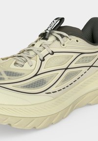 Chaussure de course beige et grise avec des panneaux en mesh, des superpositions à motifs, une semelle épaisse et rembourrée, et une languette avec une étiquette de marque.