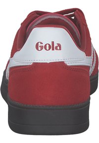 Baskets suédées rouges avec accents blancs, présentant un profil bas, un bout rond et une semelle en caoutchouc noir. Comprend un logo au niveau du talon.