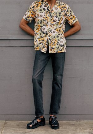 Person trägt ein gelbes und grünes, kurzärmliges Hemd mit Blumenmuster, dunkle Jeans und schwarze gewebte Sandalen und steht vor einer grauen Wand.