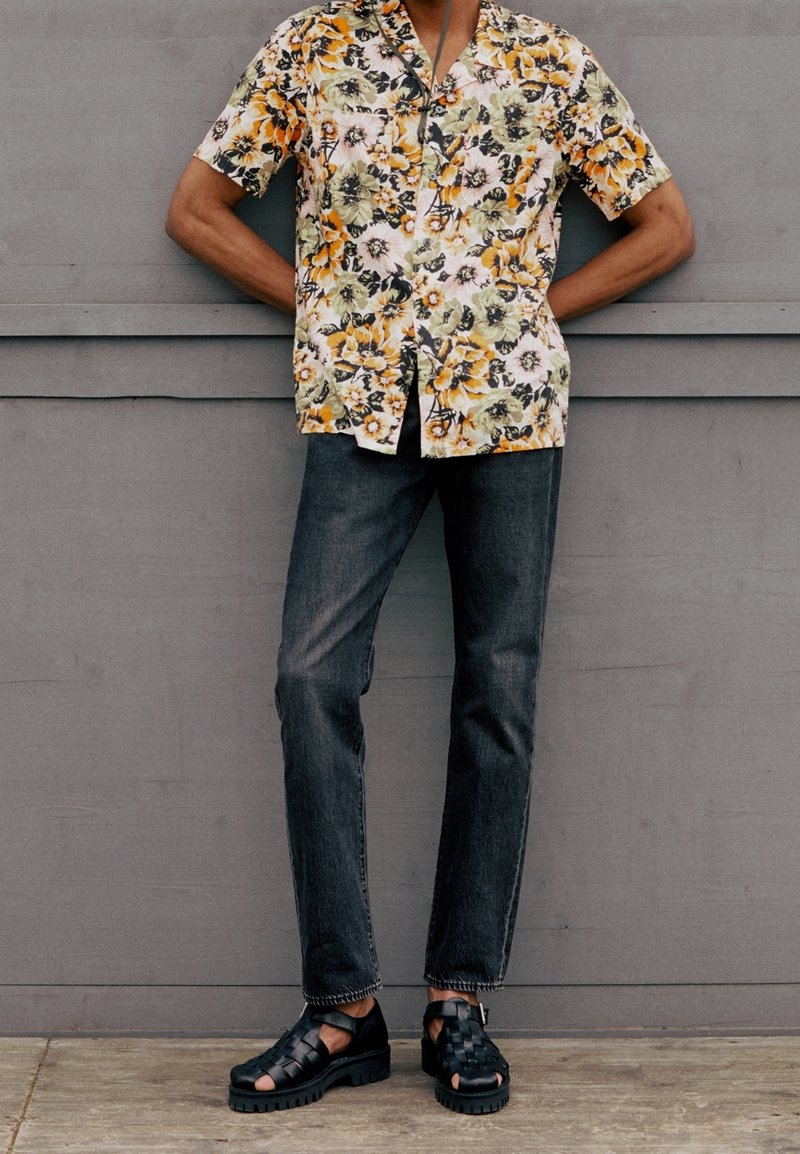 Persona vistiendo una camisa de manga corta con flores amarillas y verdes, jeans oscuros y sandalias negras entrelazadas, de pie frente a una pared gris.