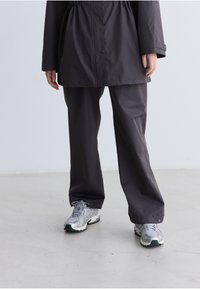 Zwarte, lichte regenjas met een verstelbare taille, gecombineerd met bijpassende broeken. De outfit wordt compleet gemaakt met zilveren sneakers met een gestructureerde zool.