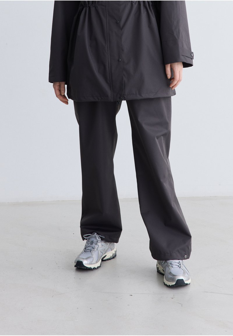 Zwarte, lichte regenjas met een verstelbare taille, gecombineerd met bijpassende broeken. De outfit wordt compleet gemaakt met zilveren sneakers met een gestructureerde zool.