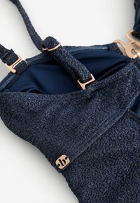 Sujetador azul marino con superficie texturizada y brillante, tirantes ajustables y detalles en oro rosa. Presenta un detalle distintivo con el logo.