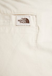 Bílá látka s hladkým povrchem, která má na zadní kapse přišitou obdélníkovou nálepku s logem "The North Face" v hnědé barvě.