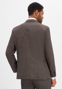 Brązowa marynarka o fakturze z wąską klapą, pojedynczym rozcięciem z tyłu i kontrastującą białą koszulą pod spodem.