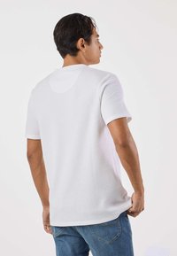 Valkoinen lyhythihainen t-paita, jossa on teksturoitu, ribbikuvioitu pintakuvio. Siinä on pyöreä kaula-aukko ja suora helma, yhdistetty sinisiin farkkuihin.