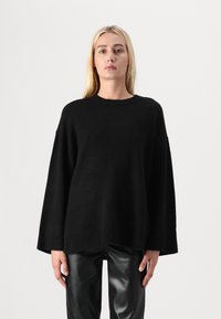 Oversized svart genser laget av mykt materiale med ribbestrikket kant og vide ermer, kombinert med skinnende svarte skinnbukser.