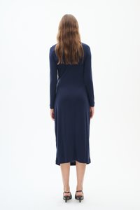 Långärmad, figurnära marinblå klänning som går till mitt på vaden, slät textur, rund nederkant, bärs med svarta högklackade skor.