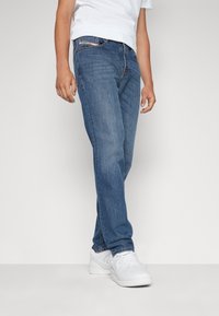 Vaqueros azules de mezclilla con un corte recto clásico, que presentan un diseño de cinco bolsillos y detalles de costura sutiles. Combinados con zapatillas blancas.