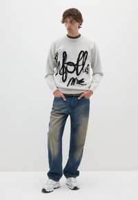 Suéter gris claro con texto negro escrito a mano, combinado con jeans azules desgastados. El modelo lleva zapatillas deportivas blancas, de pie con las manos en los bolsillos.