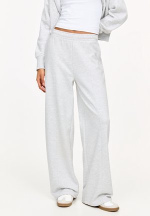 Kvinde iført løse lysegrå joggingbukser, hvid crop top, grå hættetrøje med lynlås og hvide sneakers med lysebrune såler, står med den ene hånd på hoften.