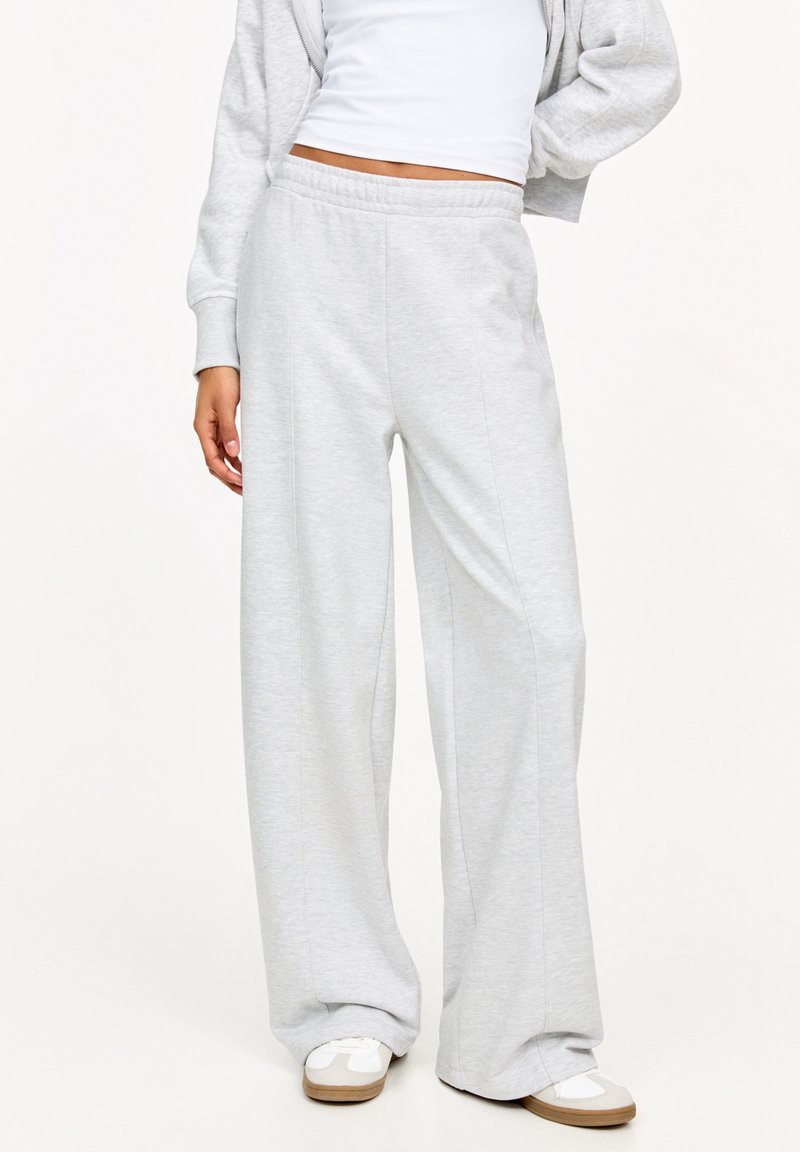 Femme portant un pantalon de survêtement ample gris clair, un crop top blanc, un sweat à capuche gris zippé et des baskets blanches avec des semelles beige, debout avec une main sur la hanche.