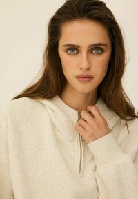 Jasnoszara bluza z kapturem na zamek błyskawiczny o teksturowanym wykończeniu. Charakteryzuje się luźnym krojem, ściągaczami przy rękawach oraz przednią kieszenią. Model trzyma zamek.