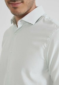 Chemise habillée vert clair, texturée, avec un col écarté, présentant des coutures subtiles et un devant boutonné.