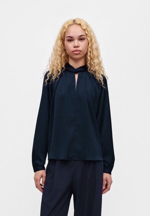 Un mannequin aux longs cheveux blonds bouclés se tient devant un fond blanc, vêtu d’un chemisier bleu marine foncé et d’un pantalon assorti.